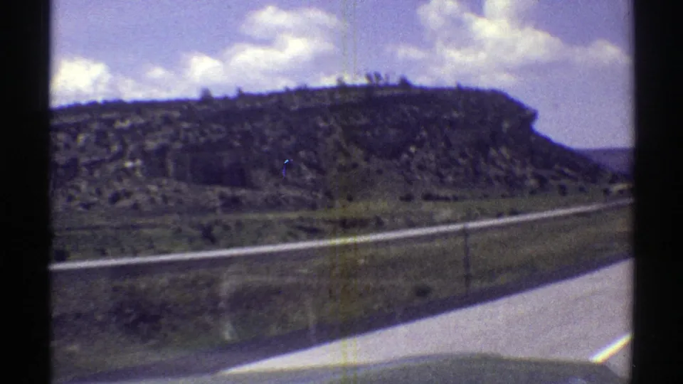 The camera passes close to a mesa slope in Arizona, with desert scrub, road edge, blue sky, and cloud cover visible.