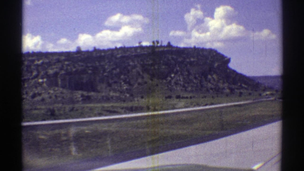 1969 Arizona 8mm view from a moving vehicle, showing a paved highway, scrubland, and eroded mesa formations under scattered clouds.