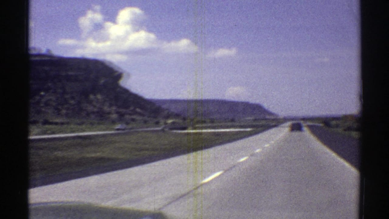 Moving-car view of an Arizona highway in 1969, with a distant vehicle, pale pavement, low vegetation, and layered rock formations.