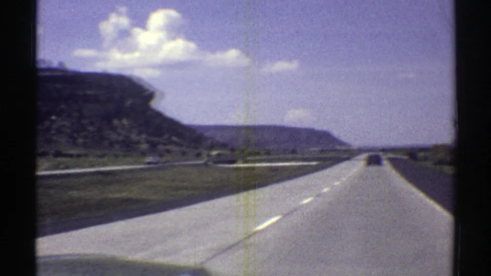 Moving-car view of an Arizona highway in 1969, with a distant vehicle, pale pavement, low vegetation, and layered rock formations.