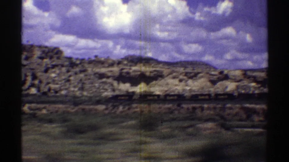 1969 Arizona desert view from a moving vehicle, with a freight train crossing the frame below eroded rock formations and clouded sky.