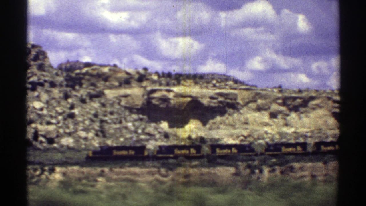 1969 Arizona 8mm frame showing Santa Fe railcars moving along tracks beneath a rugged rocky hillside and purple-tinged clouds.
