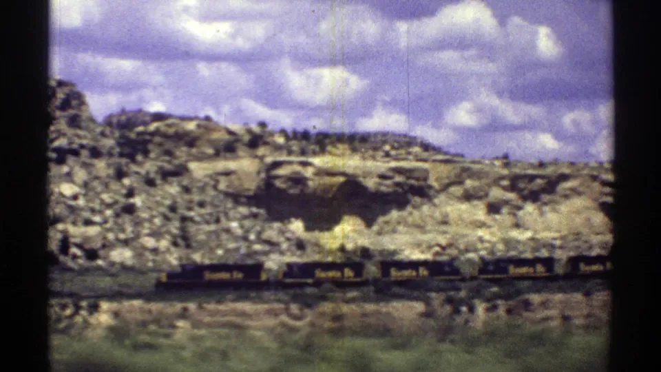 1969 Arizona 8mm frame showing Santa Fe railcars moving along tracks beneath a rugged rocky hillside and purple-tinged clouds.