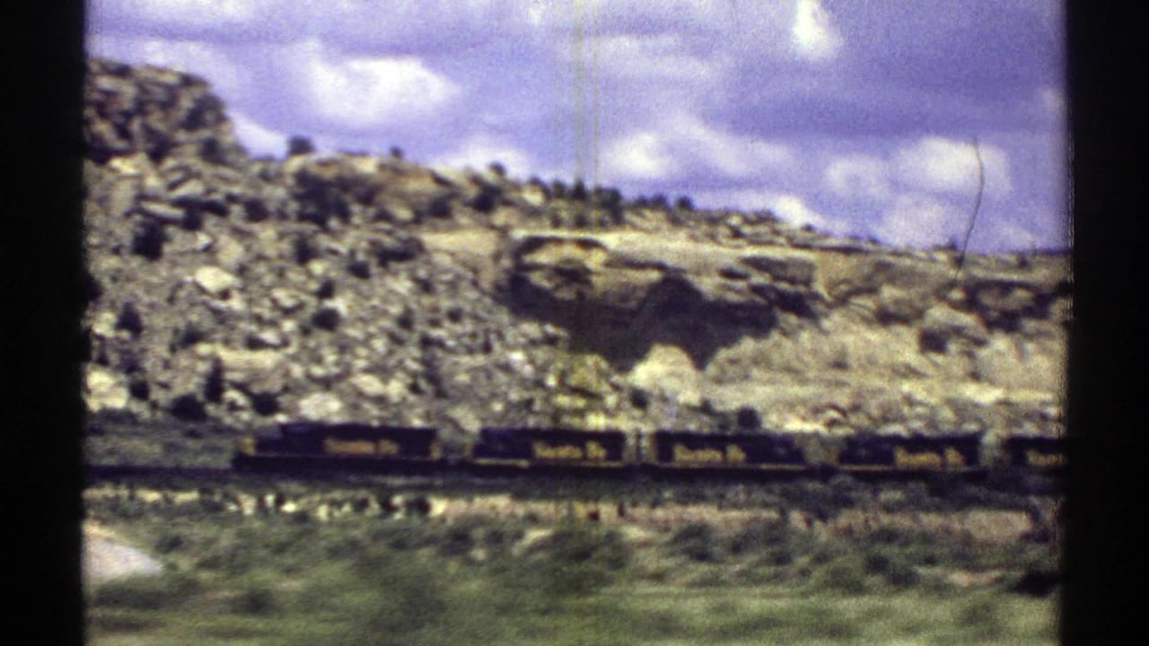 1969 Arizona 8mm frame with Santa Fe railcars passing in front of rubble slopes, pale rock ledges, and a cloud-filled sky.
