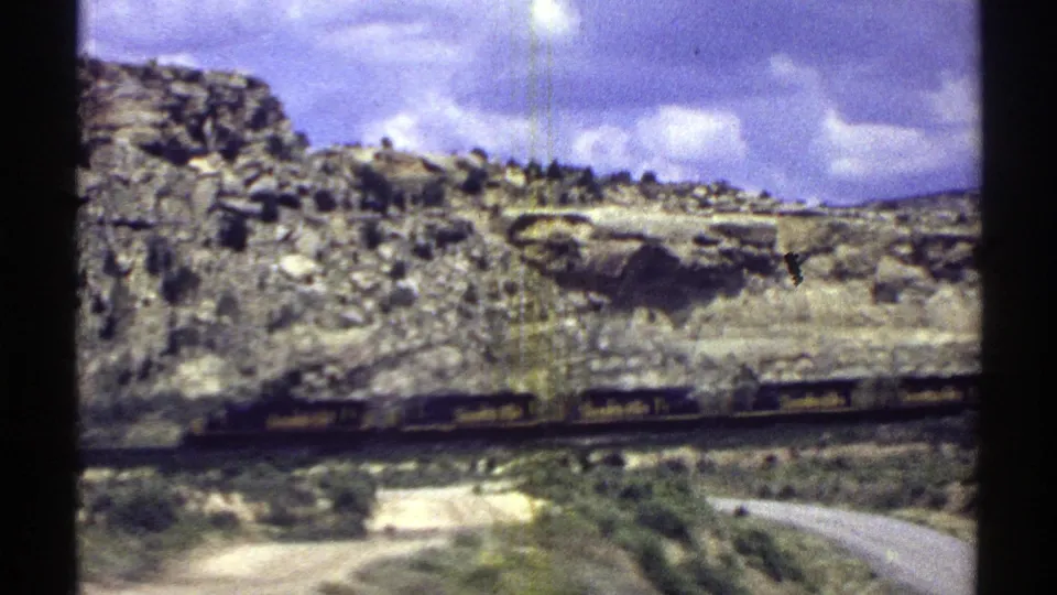 1969 Arizona 8mm frame showing a blurred Santa Fe train below rocky hills, scrub, cloudy sky, and a pale road or shoulder in the foreground.