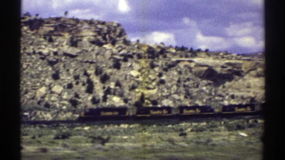 1969 Arizona 8mm frame of Santa Fe railcars along tracks beneath rugged cliffs, rock piles, sparse vegetation, and purple-tinted clouds.