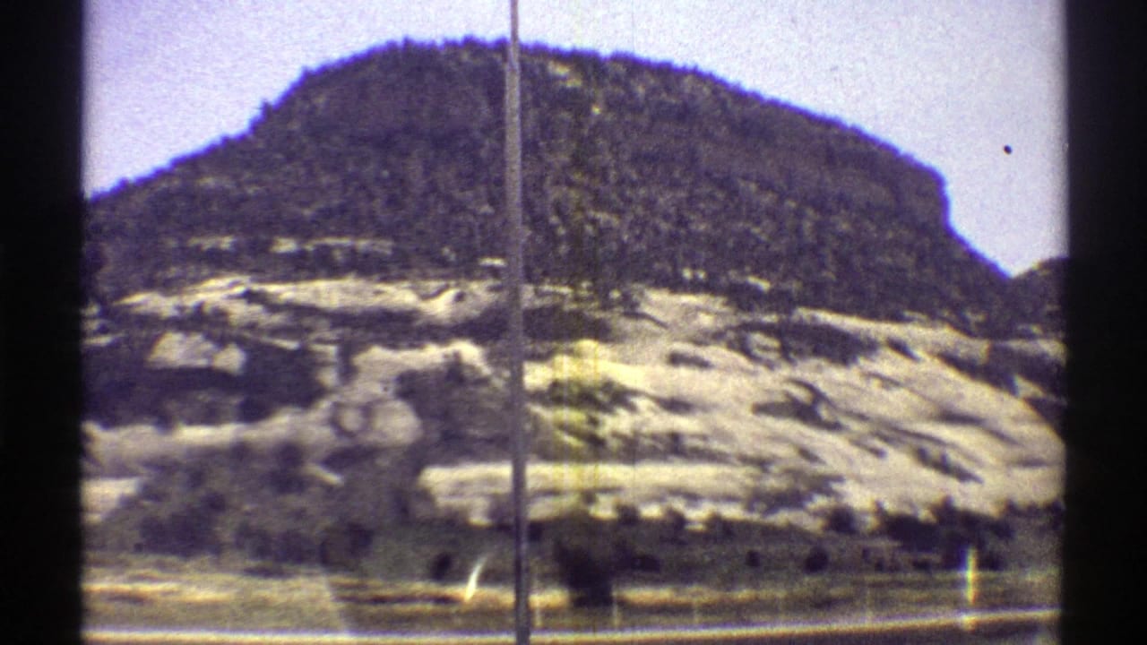 1969 Arizona 8mm frame showing a pale rocky hillside, darker mesa top, sparse vegetation, and roadside foreground from a moving car.