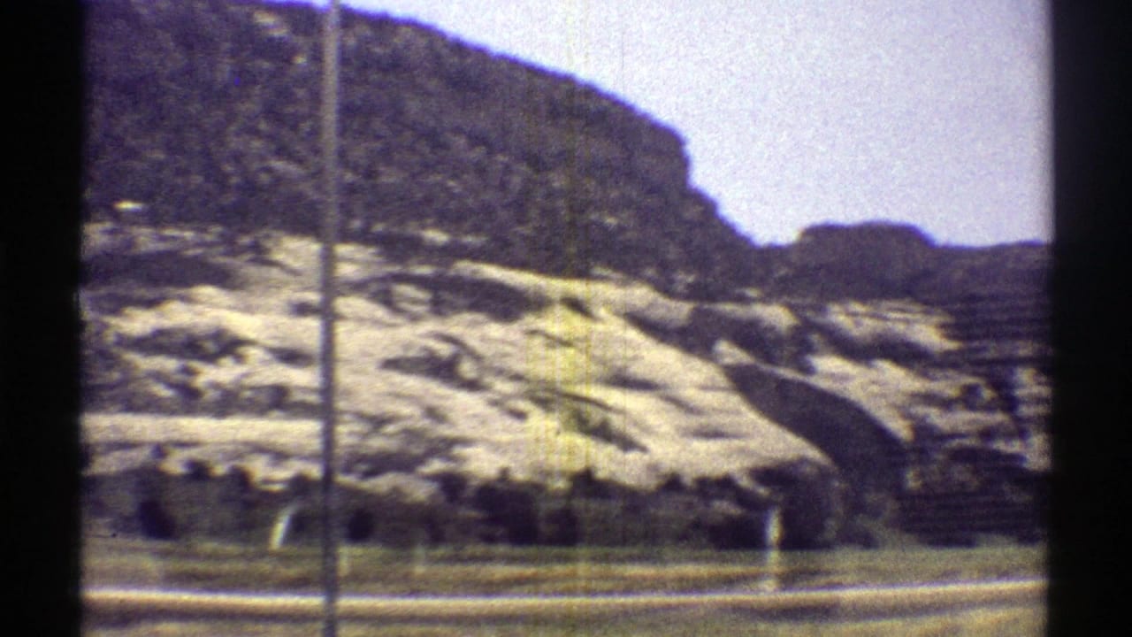 1969 Arizona 8mm frame with layered rock slopes, a darker hilltop, sparse desert plants, and roadside terrain seen in passing.