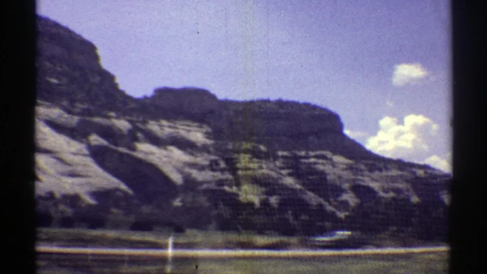 1969 Arizona home-movie frame showing low layered hills, a few bright clouds, sparse vegetation, and a roadside foreground from a car.