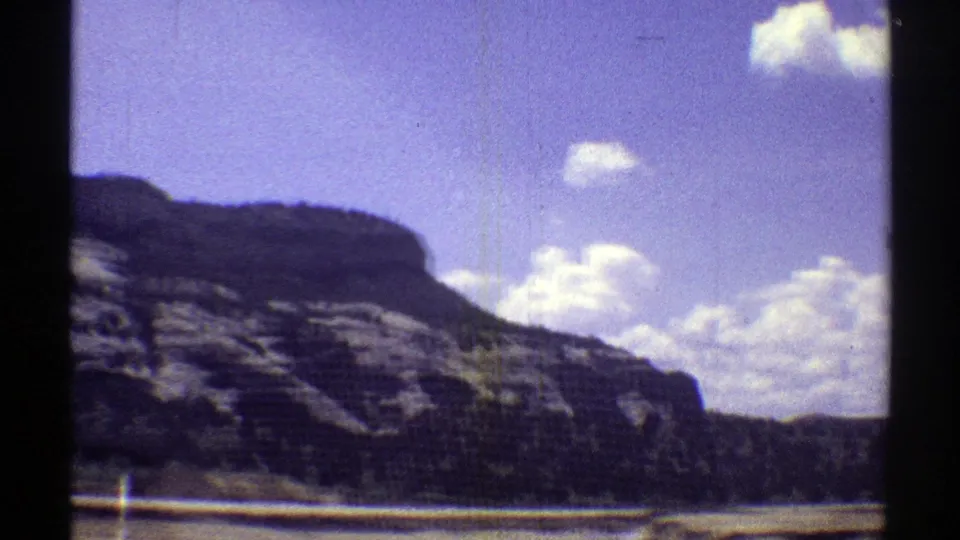 1969 Arizona 8mm frame showing a rocky ridge, sparse desert vegetation, roadside foreground, and scattered clouds viewed from a moving car.