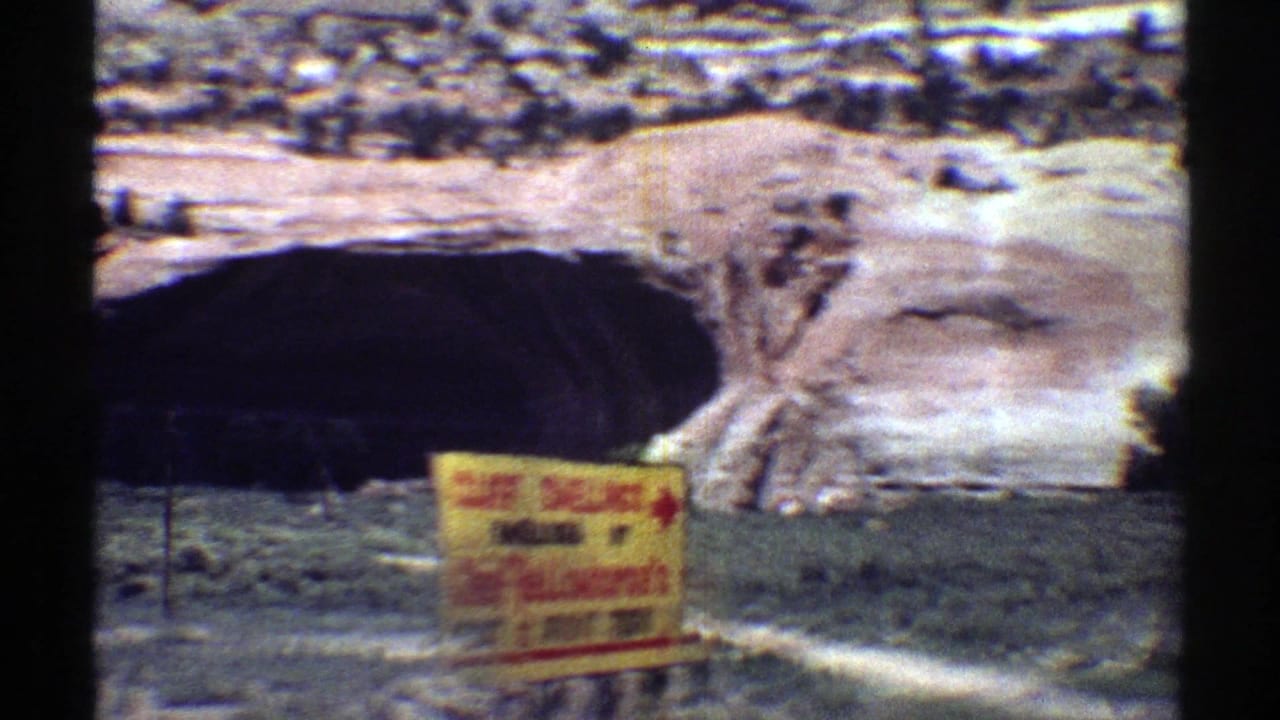 1969 Arizona 8mm frame showing a dark sandstone arch or cave opening behind roadside vegetation and a partially legible yellow sign.