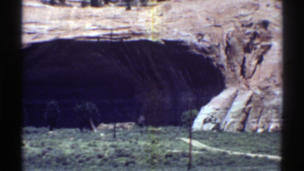 Large dark cave-like opening beneath layered sandstone cliffs and desert vegetation.