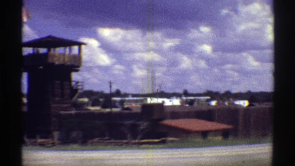 1969 Arizona home-movie frame showing a fenced roadside area, tower-like structure, distant buildings, and cloudy sky.