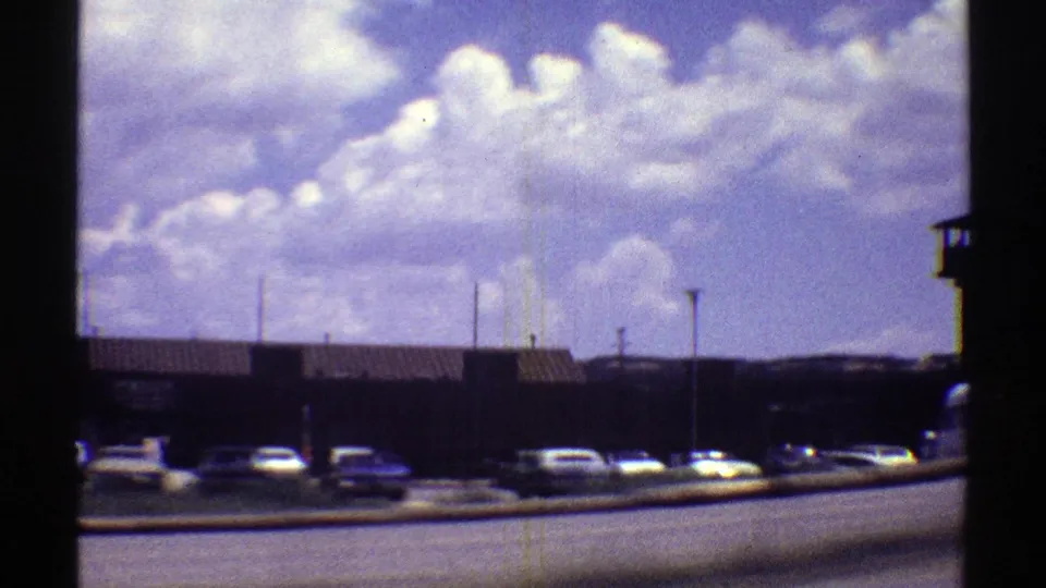 1969 Arizona 8mm frame showing low prison buildings, parked cars, fencing, and a partly cloudy sky from a roadside perspective.