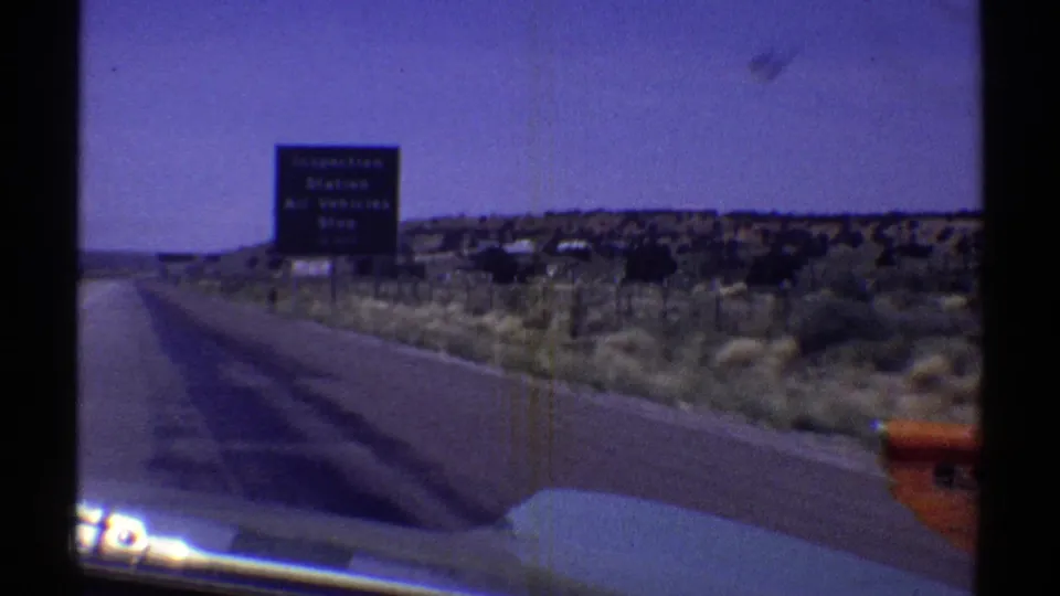 A 1969 Arizona road view shows a prison-related roadside sign, desert terrain, and part of a vehicle hood from inside a moving car.