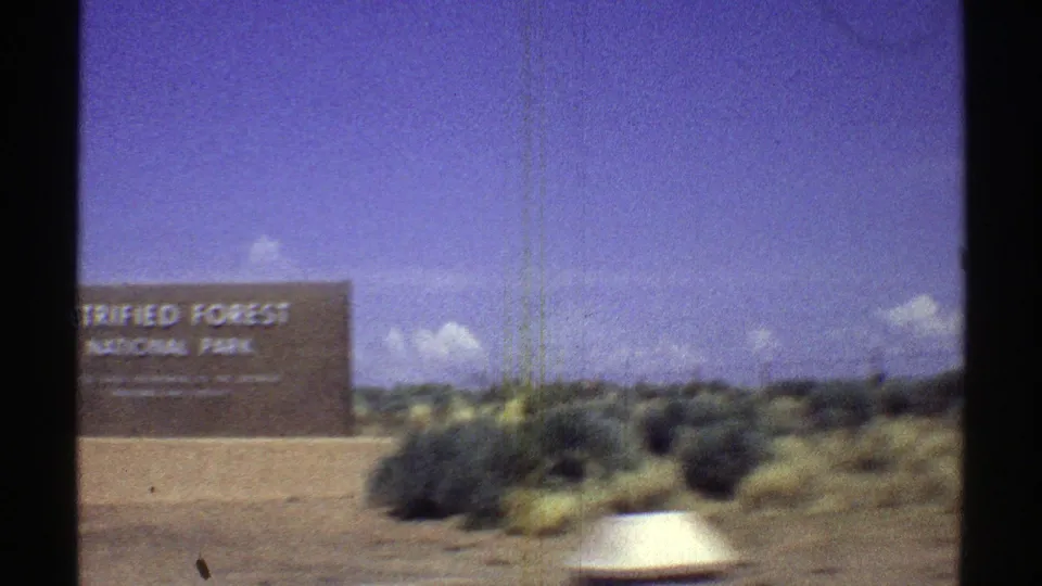 1969 Arizona road-trip frame showing the Petrified Forest National Park sign, desert vegetation, and part of a passing car.