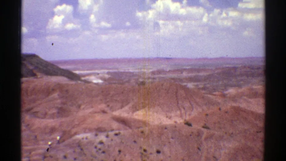 1969 Arizona 8mm frame showing reddish desert badlands, distant mesas, and a bright clouded sky near Petrified Forest National Park.