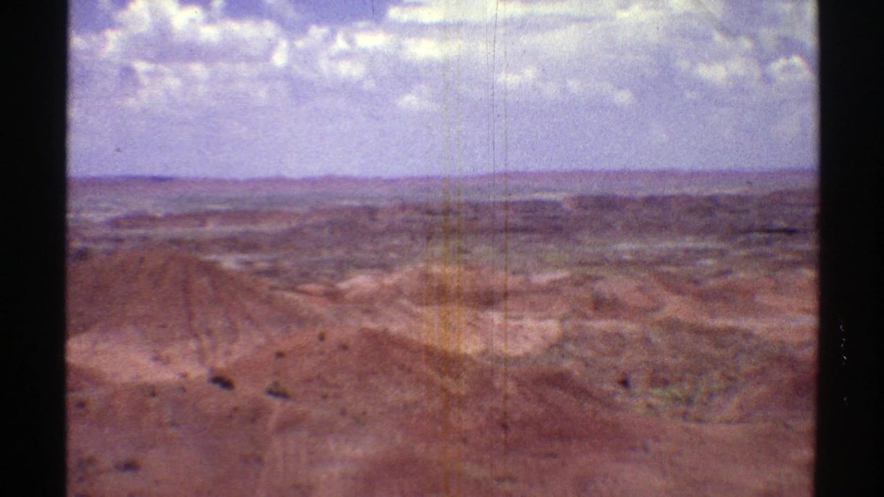 1969 Arizona 8mm frame showing eroded desert hills, sparse vegetation, and a distant horizon beneath bright clouds.