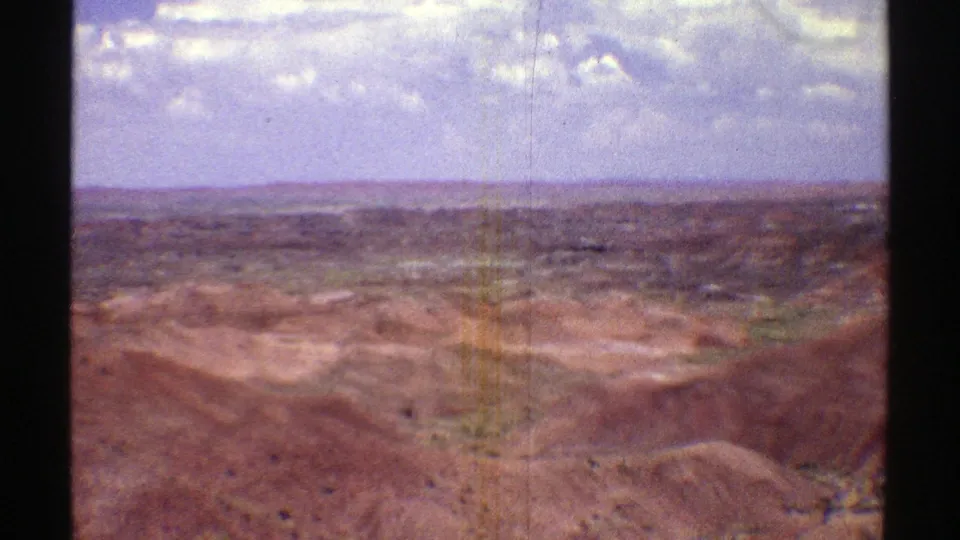 1969 Arizona home-movie frame with dry eroded ridges, open desert floor, and a pale cloudy sky during a pan.