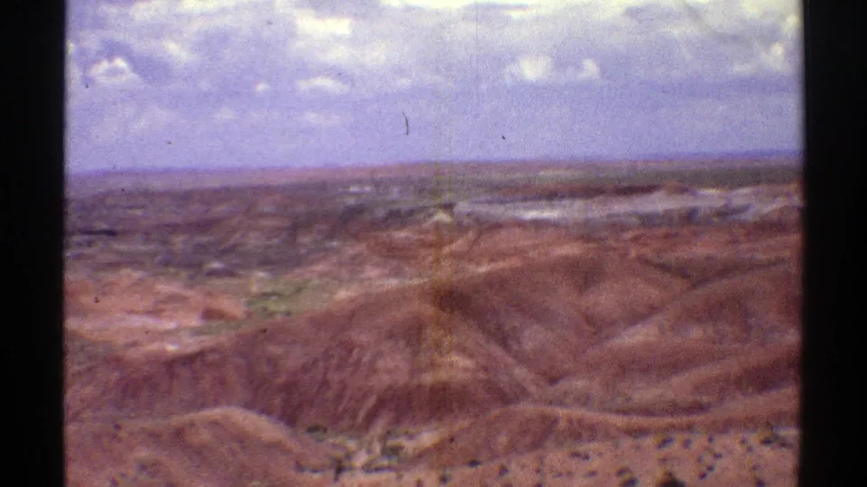 1969 Arizona 8mm frame of sunlit desert hills, sparse scrub, and a distant horizon beneath a bright clouded sky.