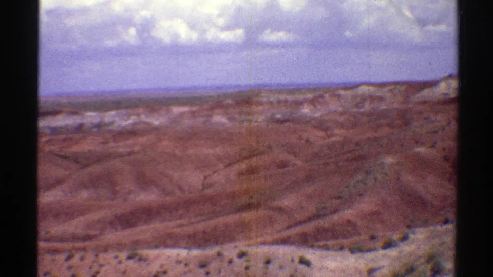 1969 home-movie view across Arizona desert hills, with a pale foreground slope, distant mesa forms, and scattered white clouds.