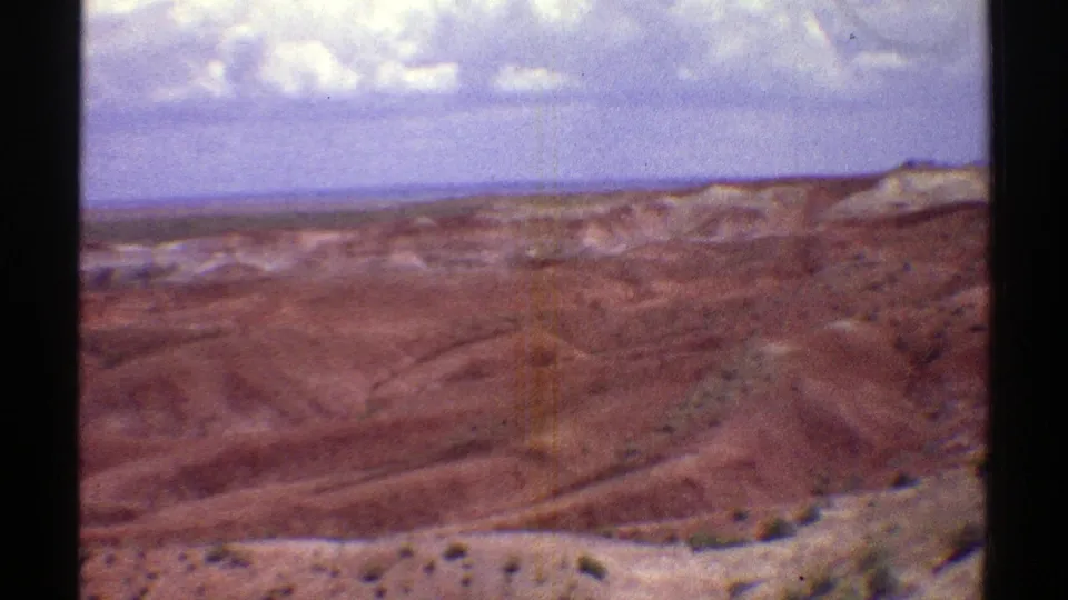 Faded 8mm frame of Arizona desert terrain, showing reddish hills, sparse scrub, distant ridges, and bright clouds overhead.