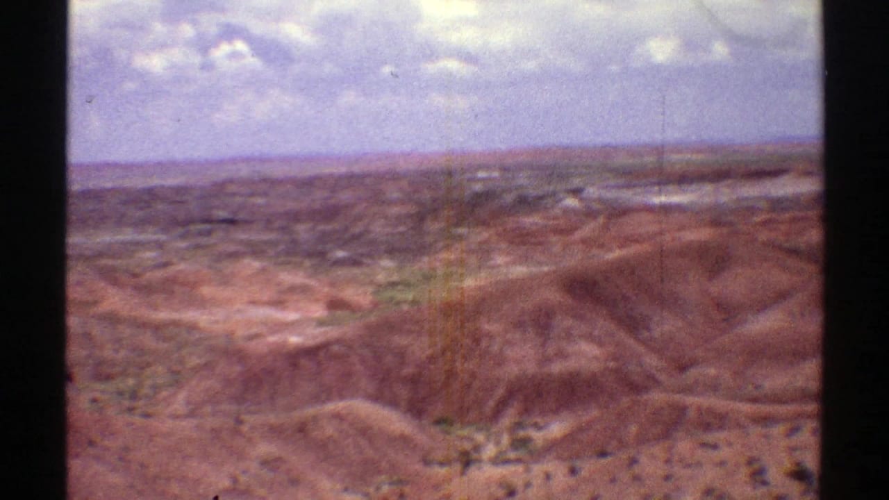 Rugged reddish-brown hills and distant desert plain under bright clouds.