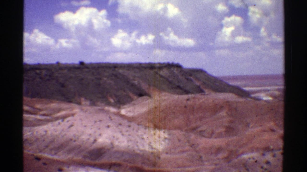 Arizona desert panorama on 8mm film, with a dark ridge, red earth slopes, and a cloud-filled blue sky.