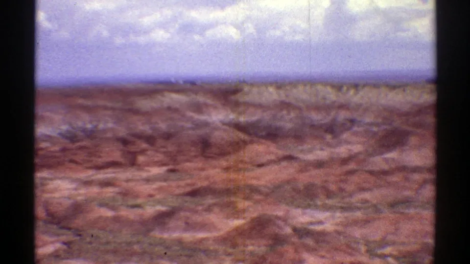 Soft 8mm frame of Arizona desert terrain, showing red-brown hills, low eroded ridges, and a hazy blue horizon.