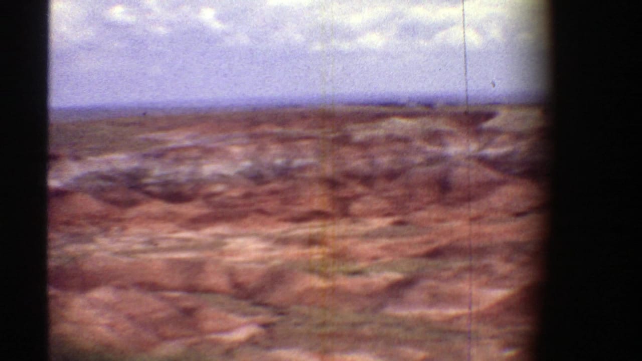 Aerial view of reddish desert ridges beneath a cloudy sky with dark film-frame edges.