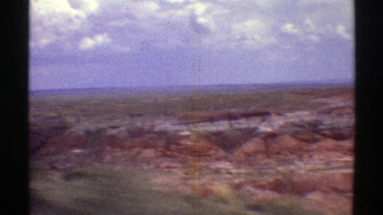 1969 Arizona home-movie frame looking across broad red and brown desert formations toward a flat horizon under clouds.