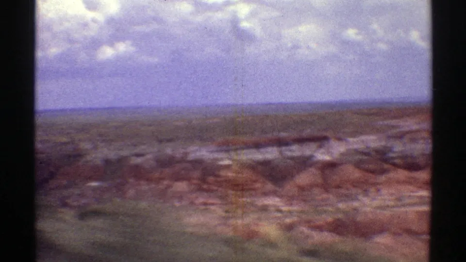 1969 Arizona home-movie frame looking across broad red and brown desert formations toward a flat horizon under clouds.