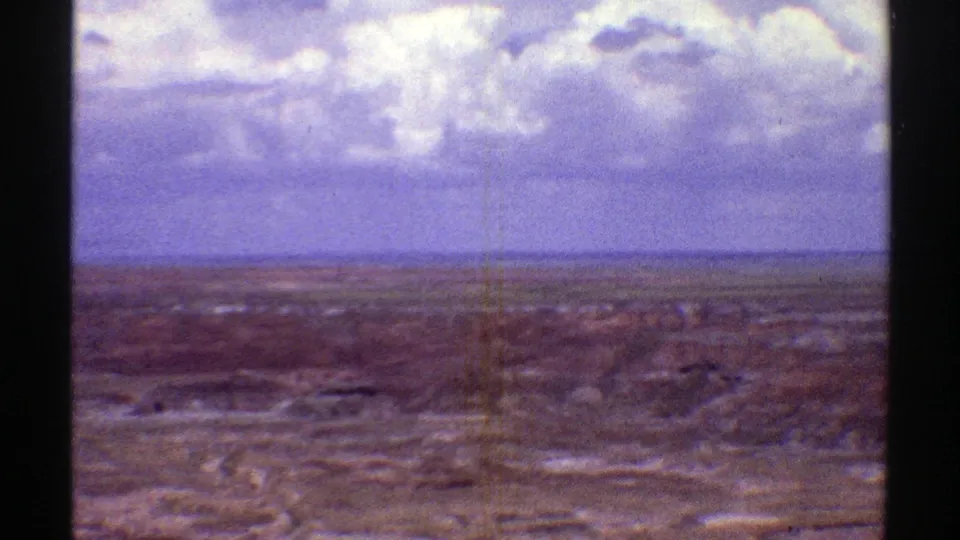 1969 8mm frame of a broad Arizona desert or field view under a cloudy sky, with soft focus and color-shifted film texture.