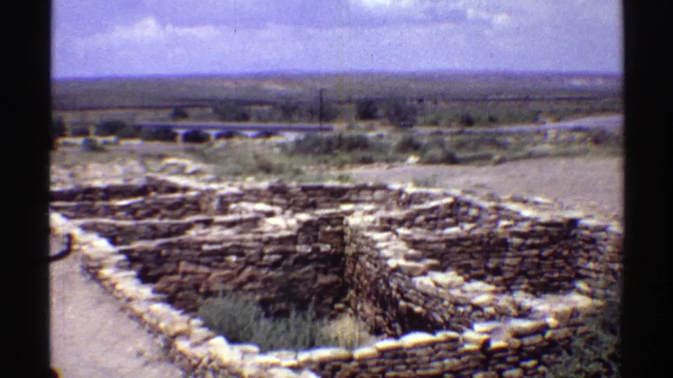 1969 Arizona home-movie frame showing low stacked stone ruins or an archaeological site in the foreground with open dry land beyond.