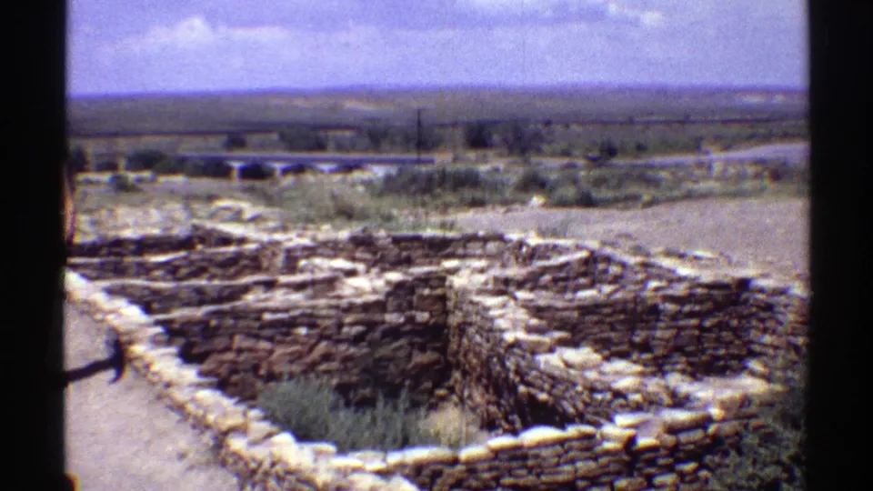 Handheld 8mm view of low stone ruins in Arizona, with dry plants in the foreground and a broad flat landscape beyond.