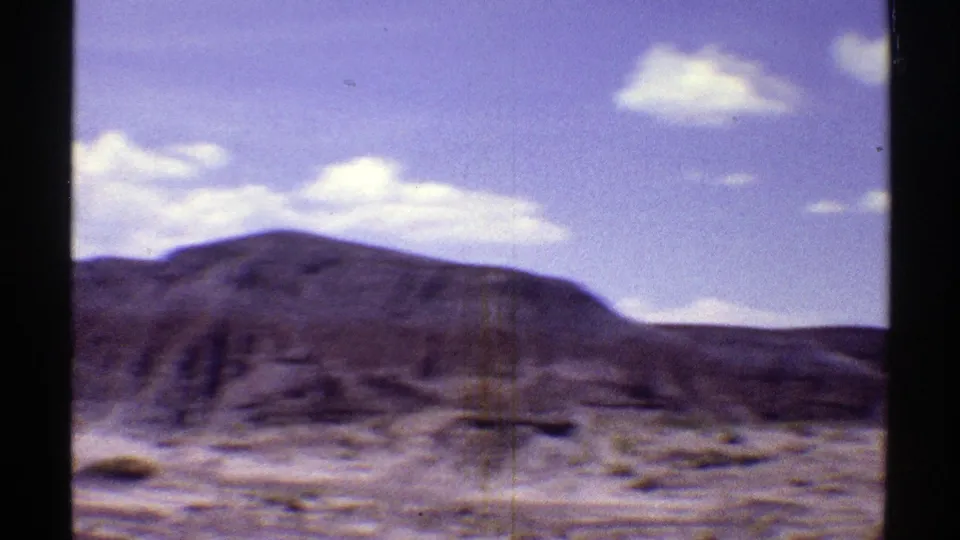 Wide 1969 Arizona home-movie view of a rocky desert ridge, scrub ground, and scattered clouds from a moving car.