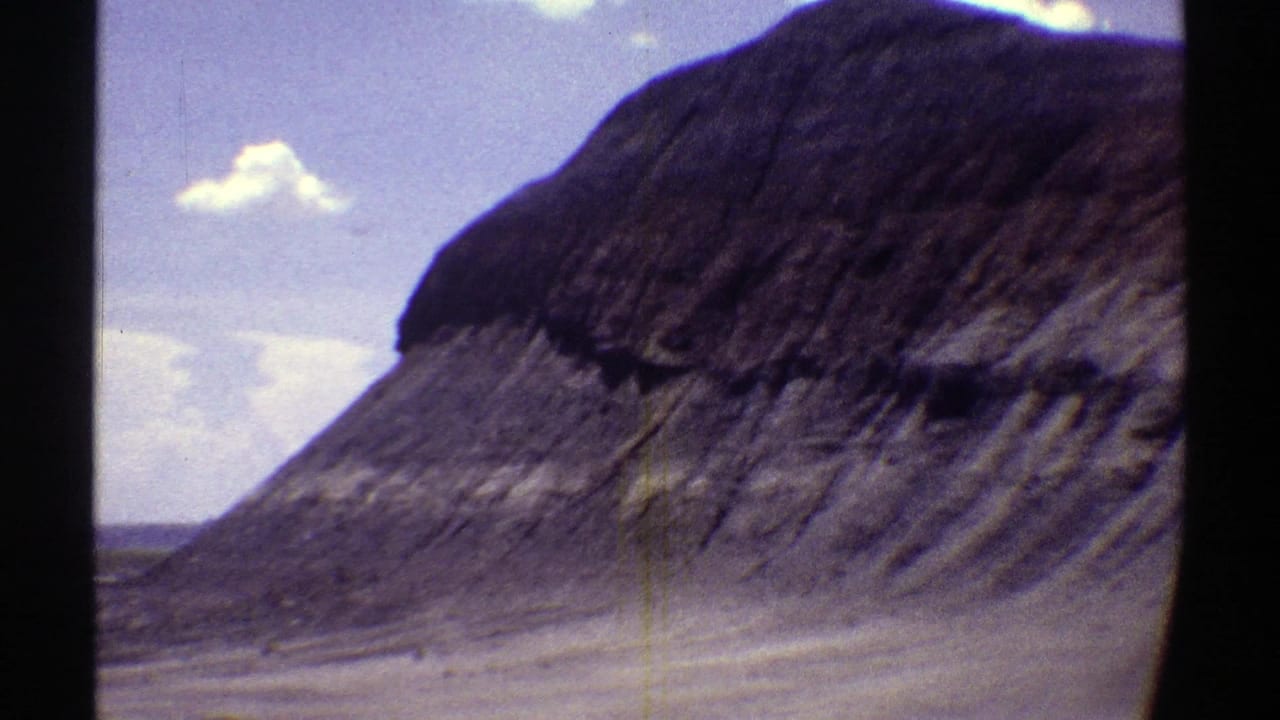 1969 Arizona 8mm frame showing a prominent layered desert butte near a wide roadway, with scrubland and scattered clouds in the background.