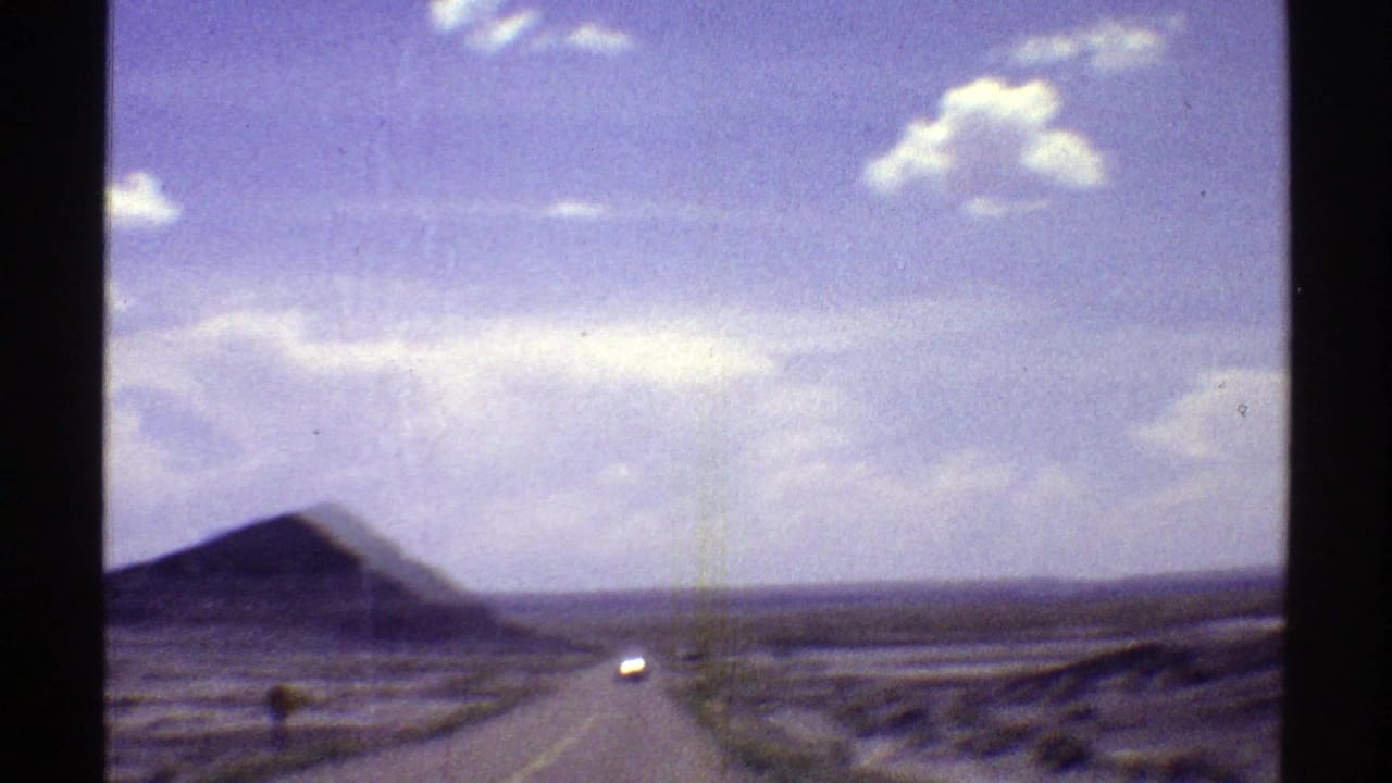 1969 Arizona home-movie view from a moving vehicle, looking down a wide straight desert road toward distant buttes and a bright sky.