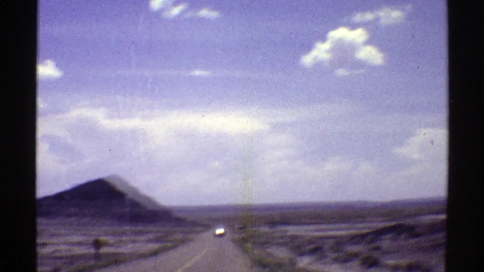 1969 Arizona home-movie view from a moving vehicle, looking down a wide straight desert road toward distant buttes and a bright sky.