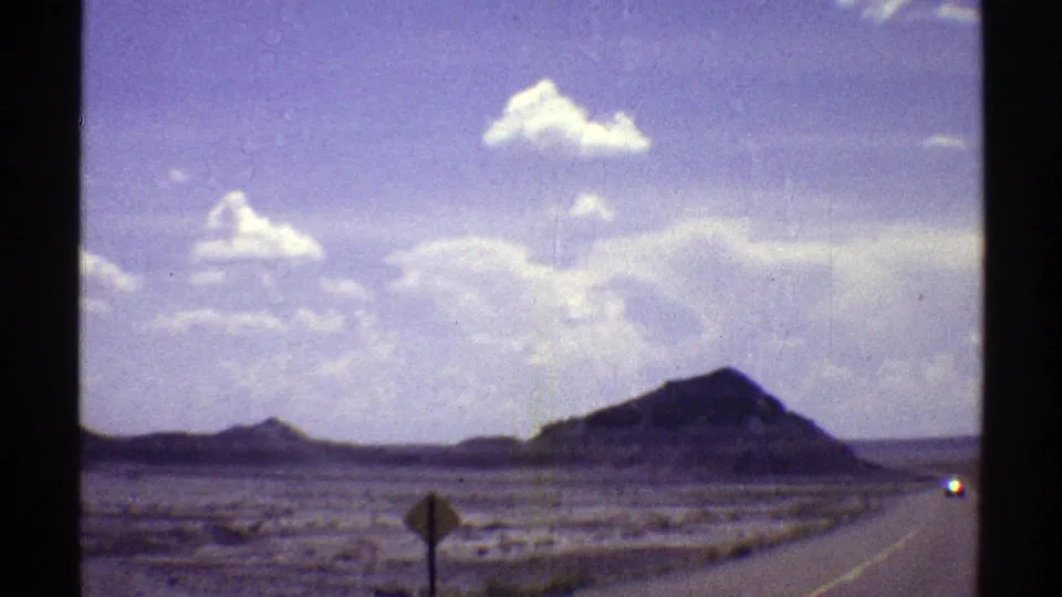 1969 Arizona 8mm frame showing a roadside sign, open highway shoulder, low desert hills, and a broad sky over arid terrain.