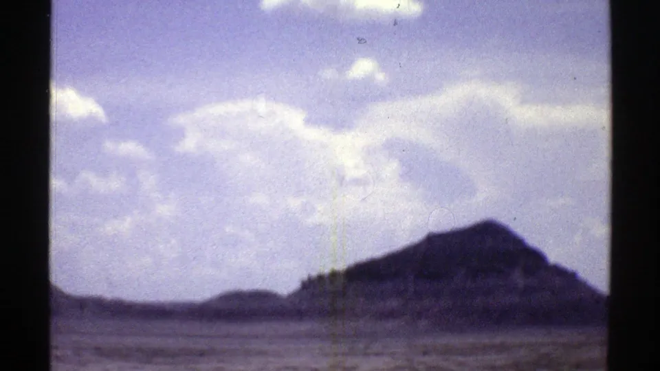 1969 Arizona home-movie frame with a dark desert butte in the distance, flat scrubland in the foreground, and bright clouds across the sky.