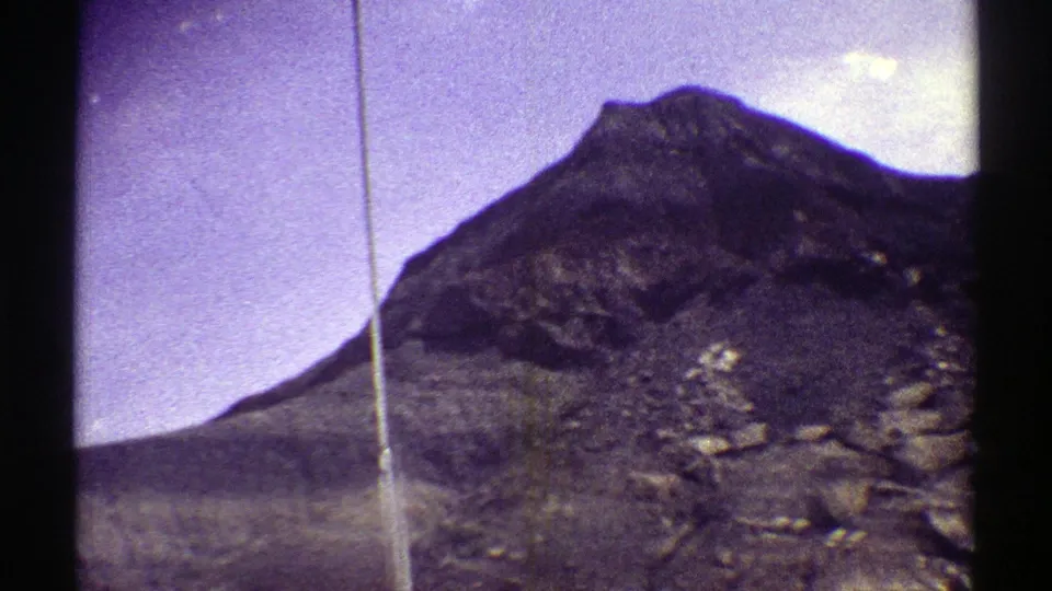 Vintage Arizona landscape frame showing a rocky peak, dry terrain, pale clouds, and prominent 8mm film wear.