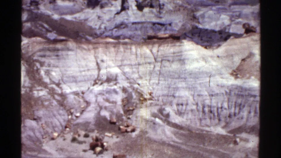 1969 Arizona desert landscape frame showing pale eroded slopes, small rocks, and a darker ridge near the horizon.