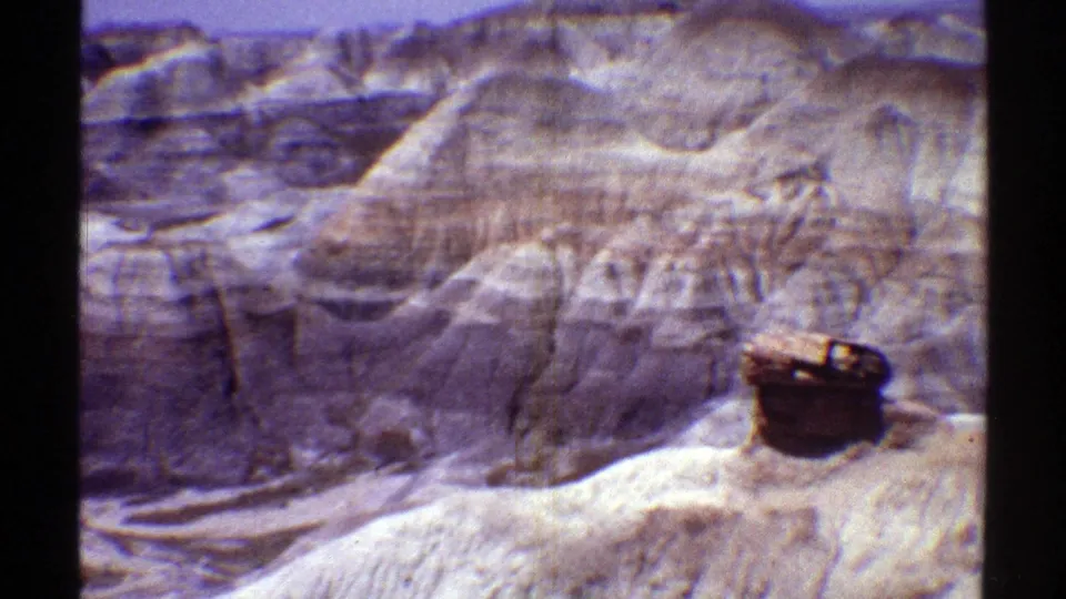 Vintage 1969 Arizona home-movie frame showing eroded cliffs, a pale ridge, and a dark barrel-like object near the lower right.