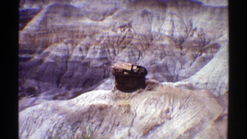 1969 Arizona 8mm frame with layered rocky formations, pale eroded ground, and a barrel-like object centered in the foreground.