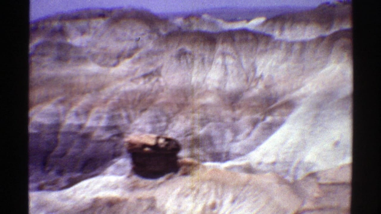 1969 Arizona 8mm frame showing layered desert badlands, pale eroded slopes, and a dark rock outcrop under a blue sky.