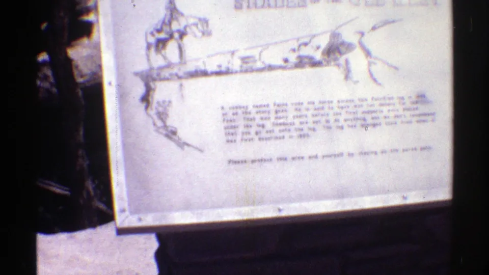 1969 Arizona 8mm frame showing a gray interpretive plaque with a cowboy drawing and text, surrounded by dark pine trunks and forest shade.