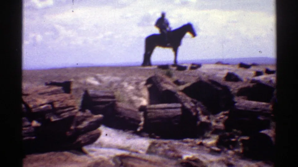 Soft 8mm frame from Arizona in 1969 showing a distant rider on a dark horse beyond weathered logs and desert rocks.