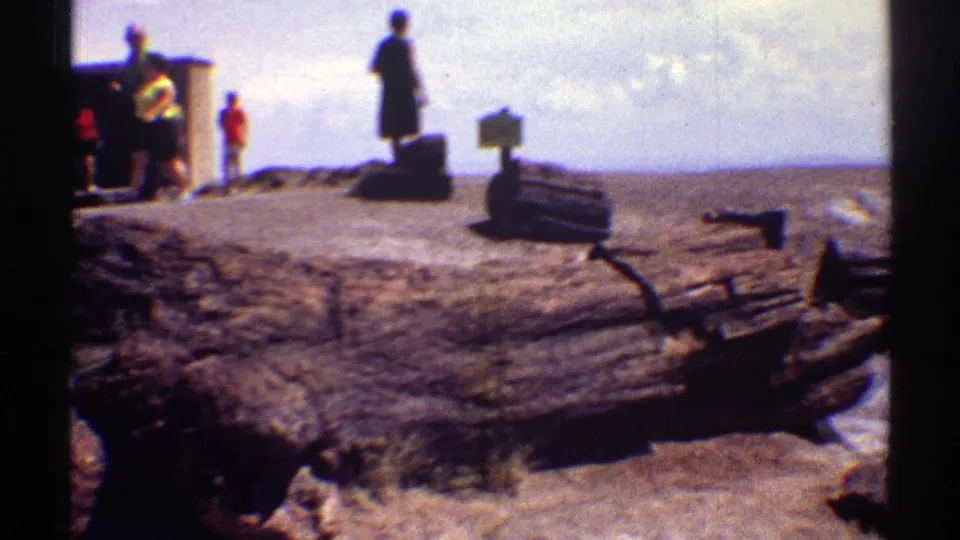 1969 Arizona 8mm frame showing distant people near a small structure or sign, with desert ground, logs, and a wide hazy sky.