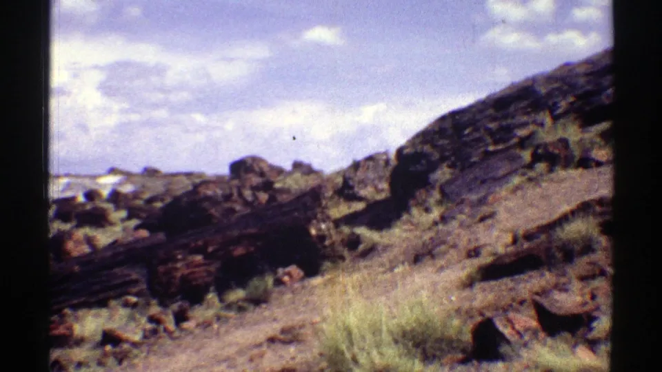 1969 Arizona 8mm frame looking across rocky arid ground with boulders, dry grass, layered stone, and clouds near the horizon.